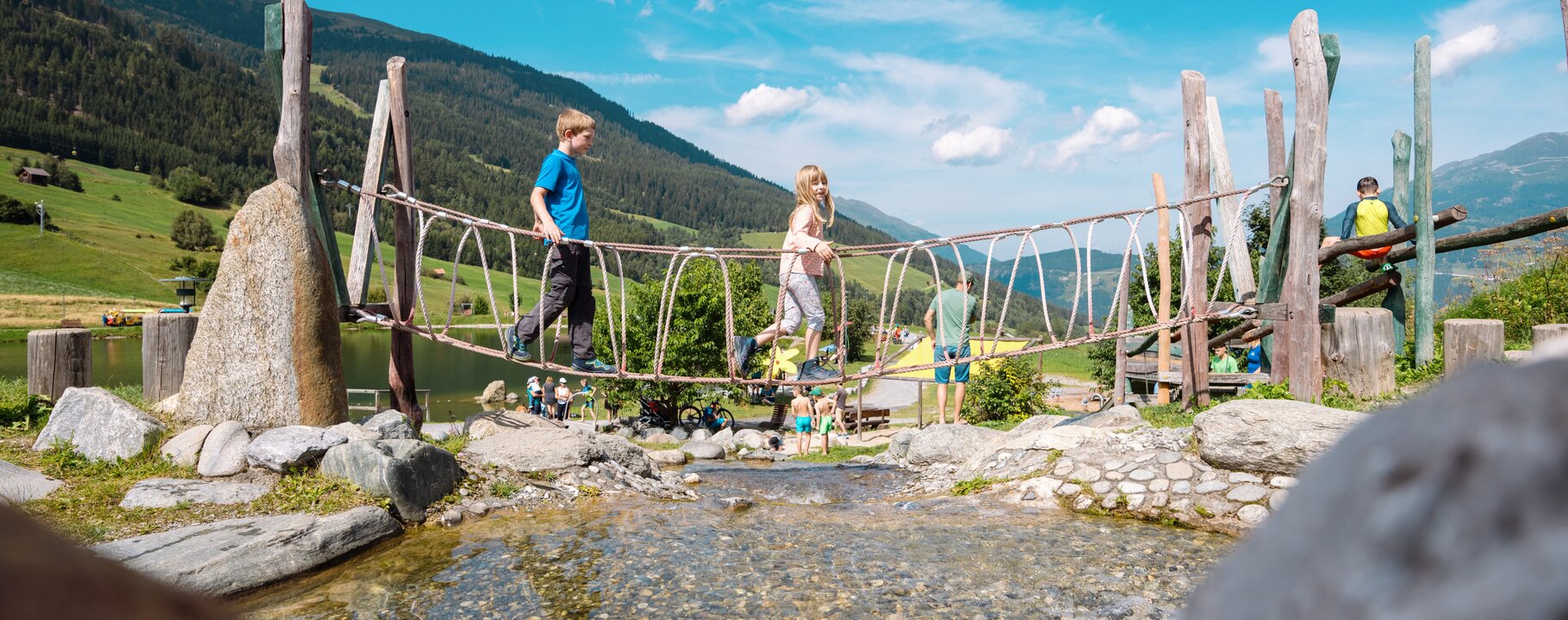 Two children balancing on a rope bridge over a stream at the Hög Adventure Park in Serfaus-Fiss-Ladis, surrounded by green meadows and mountains | © Serfaus-Fiss-Marketing GmbH | Fabian Schrigi